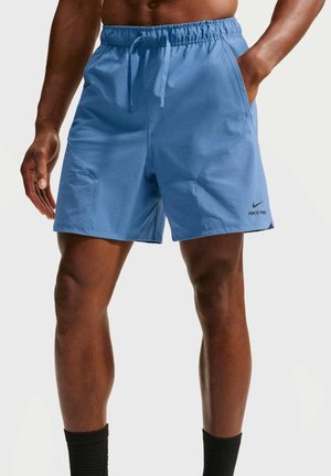 Hombre con pantalones cortos deportivos azul claro con bolsillos laterales y un logo negro de Nike Pro en la pierna izquierda, de pie con las manos parcialmente en los bolsillos.