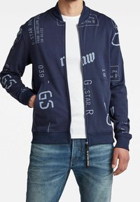 Chaqueta bomber de marina con texto gráfico en blanco y acentos de logo. Presenta un cierre de cremallera en la parte delantera, puños de canalé y dos bolsillos laterales. Material de mezcla de algodón.