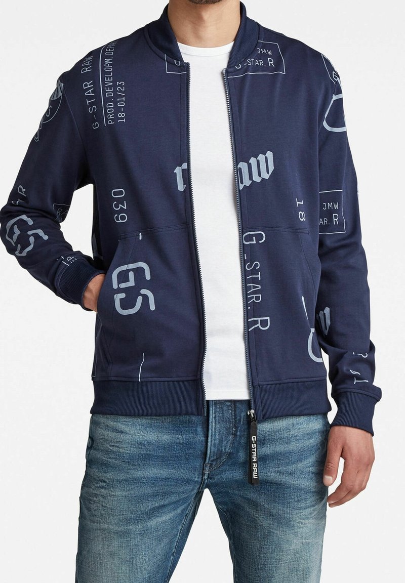 Chaqueta bomber de marina con texto gráfico en blanco y acentos de logo. Presenta un cierre de cremallera en la parte delantera, puños de canalé y dos bolsillos laterales. Material de mezcla de algodón.