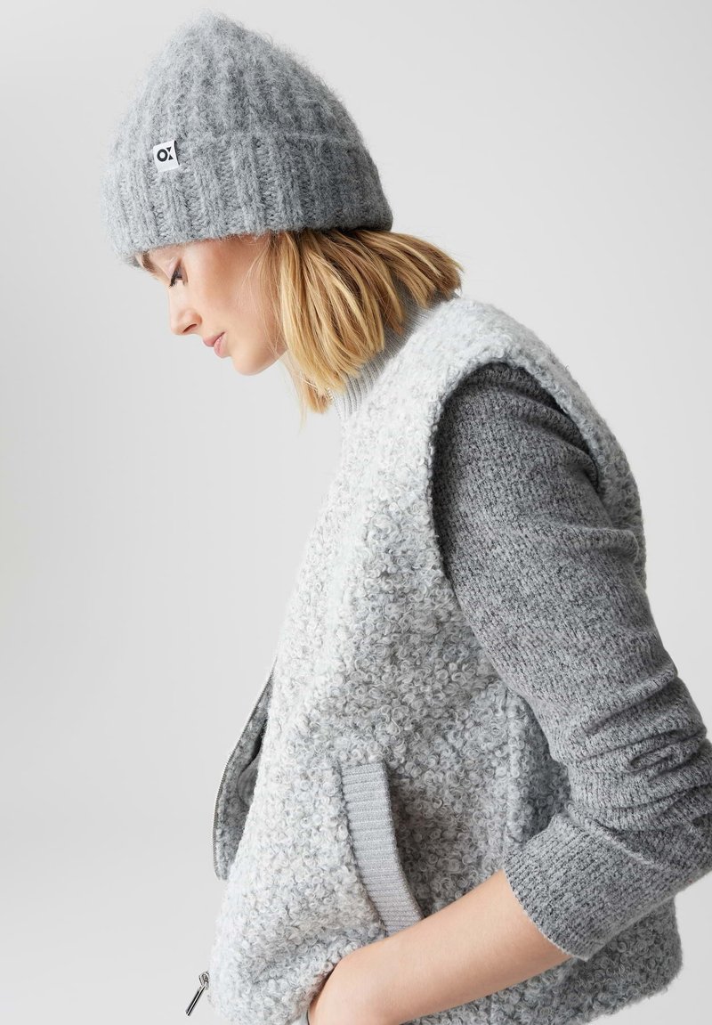 Grijze gebreide beanie met een label, gecombineerd met een textuurgrijze mouwloze vest en een lange mouw gebreid top, die gelaagde grijstinten toont.