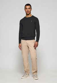 Jersey de punto gris oscuro con mangas largas, cuello redondo y un pequeño emblema en el pecho, combinado con pantalones chinos beige y zapatillas claras.