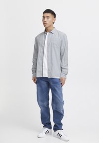 Camicia button-up a righe grigie sopra una maglietta bianca, abbinata a jeans blu e sneakers bianche con strisce nere. Vestibilità slim e design casual.