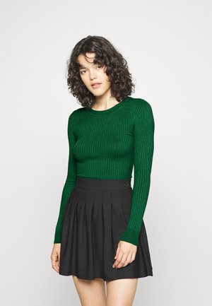 Maglione verde a costine con maniche lunghe abbinato a una gonna nera plissè. Il maglione presenta un collo rotondo e un design aderente.