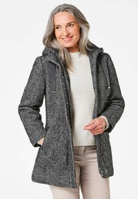 Haringbone-patroon wollen jas in zwart en wit, met een capuchon, zijzakken en grijze accenten. Zachte textuur past goed bij een lichte trui.