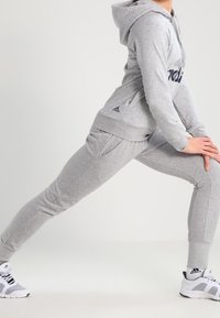 Sudadera gris con capucha y bolsillo tipo canguro, combinada con pantalones deportivos grises a juego. Presenta zapatillas deportivas con acentos negros.