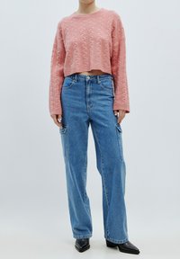 Rosa Kabelstrick-Cropped-Sweater mit langen Ärmeln, kombiniert mit blauen, lässig geschnittenen Cargo-Jeans und schwarzen, spitzen Stiefeletten.