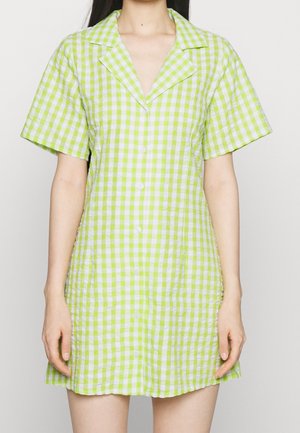 Kobieta w krótkiej sukience zapinanej na guziki z krótkim rękawem w zielono-białą kratkę, stojąca na jednolitym jasnym tle.