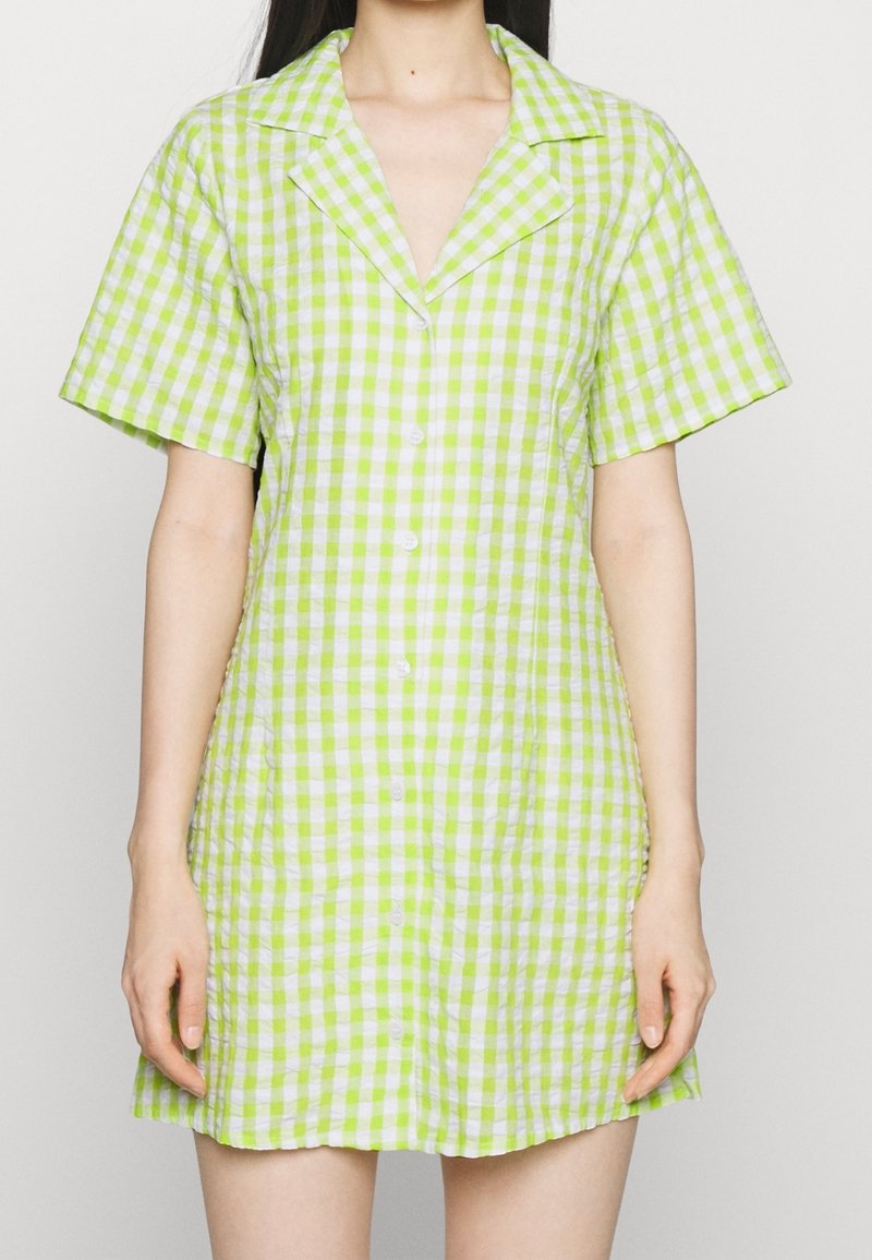 Femme portant une robe mini boutonnée à manches courtes avec un motif vichy vert et blanc, debout contre un fond clair uni.