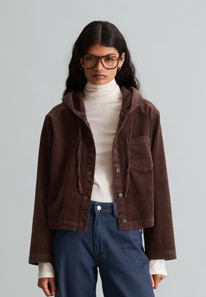 Femme aux longs cheveux foncés portant de grandes lunettes, un col roulé blanc, une veste en velours côtelé marron et un pantalon bleu taille haute, debout sur un fond gris.