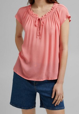 Frau trägt eine rosa Bluse mit kurzen Ärmeln, gerafftem Ausschnitt und Schnürdetail, kombiniert mit dunkelblauen Jeansshorts.