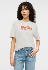 Ανοιχτό γκρι κοντομάνικο T-shirt με κόκκινο μοτίβο καρδιάς και τη λέξη "HUGO" στο κέντρο. Συνδυάζεται με ανοιχτό μπλε τζιν και ζώνη.