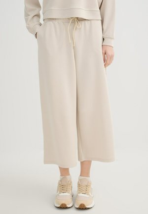 Pantalones cortos de pierna ancha beige con cintura de cordón, combinados con sudadera beige a juego y zapatillas deportivas sobre un fondo liso.