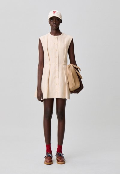 Vestido de lino beige con botones y plisados, combinado con un bolso beige y zapatos marrones con cordones azules. Calcetines rojos completan el conjunto.