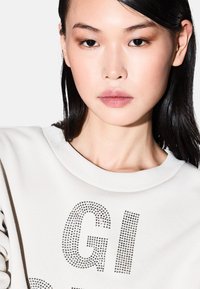 Sudadera blanca con tejido texturizado, presenta letras de strass "GI" y detalle de mangas con volantes, cuello redondo. Modelo con cabello negro lacio.
