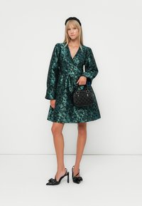 Woman wearing a green floral patterned dress, black headband, black high-heeled slingback shoes, and holding a small black handbag.