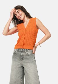 Gilet tricoté orange avec un décolleté en V et sans manches, présentant un motif texturé, associé à un jean gris taille haute.