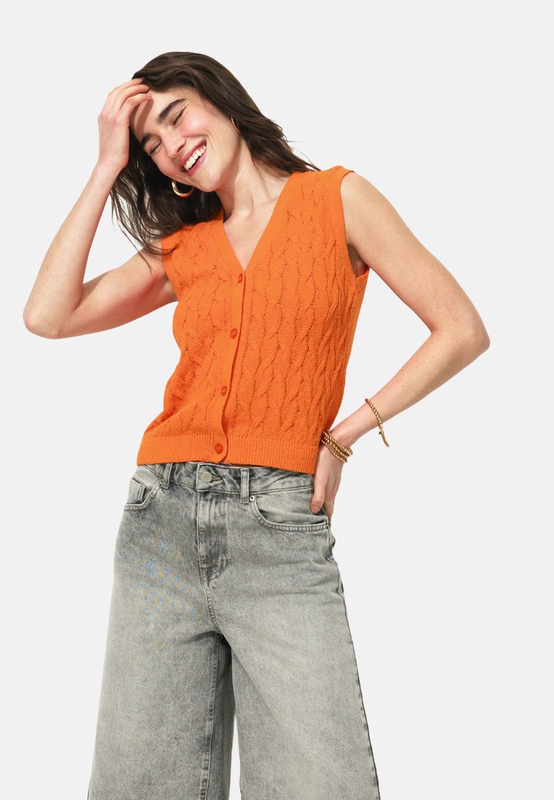 Gilet tricoté orange avec un décolleté en V et sans manches, présentant un motif texturé, associé à un jean gris taille haute.