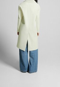 Ljusgrön, lång ullkappa med bred krage, bakventil och rak silhuett, kombinerad med lösa blå denimbyxor.