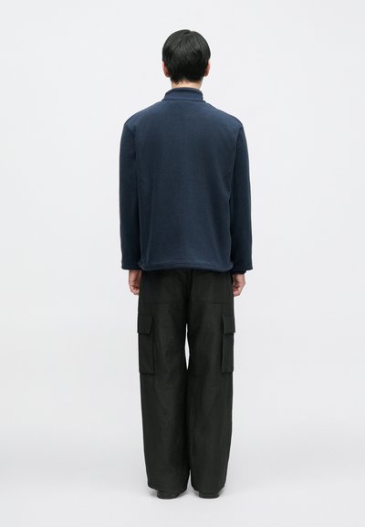 Tummansininen yläosa, jossa on korkea kaulus ja pitkät hihat, yhdistettynä väljiin mustiin cargo-housuihin, joissa on paikkataskut.