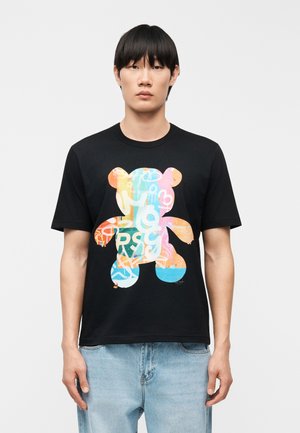 Νέος άνδρας που φοράει μαύρο μπλουζάκι με πολύχρωμο γραφικό αρκουδάκι γεμάτο αφηρημένα σχήματα και γράμματα, συνδυασμένο με ανοιχτόχρωμα τζιν παντελόνια.
