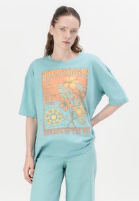 Camiseta oversized turquesa con gráfica floral y texto. Mangas cortas, corte relajado, material de algodón suave, combinada con pantalones a juego.