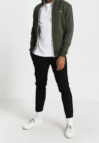 Man med olivgrön zip-up jacka, vit T-shirt, svarta joggerbyxor, vita strumpor och vita sneakers, stående med händerna i fickorna.