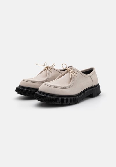 Beige Wildlederschuhe mit runder Zehenpartie, farblich passendem Nähen und schwarzer Gummisohle mit geriffelter Textur, kombiniert mit dünnen Schnürsenkeln.
