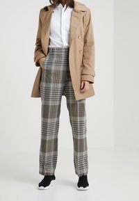 Person som bär beige trenchcoat, vit skjorta, högmidjade rutig byxor och svarta sneakers med vita sulor, står med ena handen i fickan.