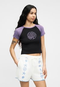 Γυναίκα με μακριά σκούρα μαλλιά που φοράει μαύρο crop top με μωβ μανίκια και σχέδιο ήλιου, συνδυασμένο με λευκά σορτς που έχουν μπλε σχέδια με θέμα τη θάλασσα.