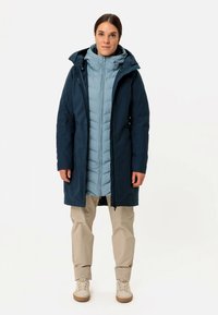 Tumši zils ūdensnecaurlaidīgs virsjaka pār gaiši zilu pūkainu jaku, valkāts kopā ar bēšām biksēm un krēmkrāsas sporta apaviem. Aprīkots ar kapuci un priekšējo rāvējslēdzēju.