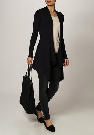 Femme portant un long cardigan noir, un top beige, un pantalon skinny noir et des ballerines noires, tenant un grand sac à main noir dans sa main gauche.