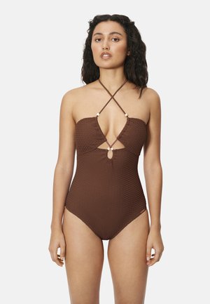 Mujer con un traje de baño entero marrón texturizado, con tirantes cruzados en forma de halter y un recorte en forma de ojo de cerradura en el busto, de pie frente a un fondo liso.