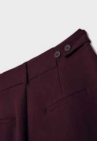 Detailný záber tmavohnedých nohavíc ukazujúci pás s dvoma gombíkmi, pätky na opasok a zadné vrecká s lepením na neutrálnom pozadí.