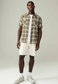 Korte mouwen, geruite shirt in beige en groentinten, met twee voorzakken, gecombineerd met crèmekleurige shorts en lichtgrijze sneakers.