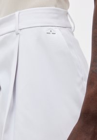 Λευκό παντελόνι με ψηλή μέση, θηλιά για ζώνη και μικρό μεταλλικό λογότυπο στο πλάι, φορεμένο από άτομο με σκούρο δέρμα και μπεζ τοπ.