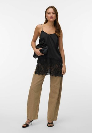 Femme portant un caraco noir bordé de dentelle, un pantalon beige à jambes larges, des sandales noires à talons, tenant une pochette noire.