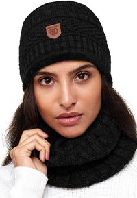 Czarna dziana czapka beanie z prążkowanym wzorem i skórzaną metką logo, zestawiona z pasującym, przytulnym czarnym szalikiem, noszona na białym swetrze.