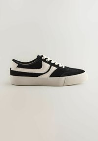 Musta-valkoinen mokkasneakeri, jossa on matala malli, pyöreä kärki ja valkoiset nauhat. Varustettu teksturoidulla pohjalla ja erottuvalla ompelulla.