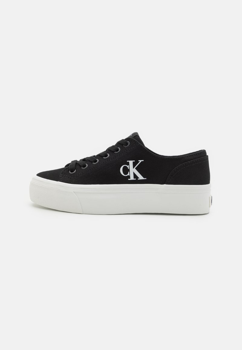 Tenisi negri din pânză cu talpă și orificii din cauciuc alb. Prezintă un logo cusut "cK" pe partea exterioară. Design low-top cu vârf rotund.