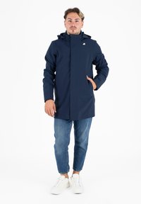 Cappotto impermeabile navy con cappuccio, chiusura frontale a zip e tasche laterali. Indossato sopra a jeans blu casual e sneakers bianche. Tessuto dalla superficie liscia.
