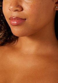 Gros plan sur un cou et un visage lisses de couleur brun clair avec des taches de rousseur visibles, des lèvres brillantes et une petite boucle d'oreille argentée. La peau a un éclat subtil.