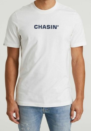 Homme portant un T-shirt blanc à manches courtes avec « CHASIN' » en lettres bleues marine en gras, associé à un jean bleu clair usé.