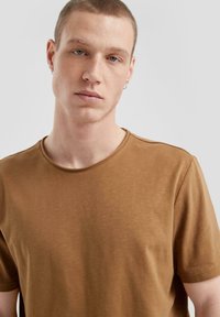 Brązowa bawełniana koszulka z okrągłym dekoltem i krótkimi rękawami. Charakteryzuje się gładką fakturą i subtelnymi detalami przeszycia wzdłuż krawędzi.