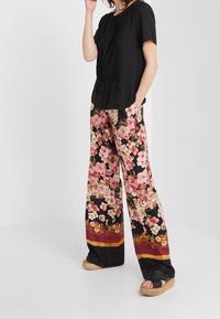 Haut noir à plis, associé à un pantalon large à fleurs roses et crème sur fond noir, agrémenté d'une bande d'accent bordeaux et or.