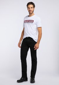 Biała koszulka z napisem "MUSTANG TRUE DENIM BRAND" w burgundzie, w połączeniu z czarnymi dżinsami i czarnymi butami, stojąca na jasnym tle.