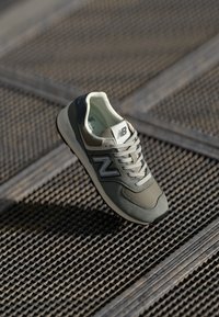 Sneaker New Balance grigia e beige con lacci posizionata su una superficie a griglia metallica in un'illuminazione soffusa.