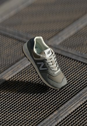 Sneaker New Balance grigia e beige con lacci posizionata su una superficie a griglia metallica in un'illuminazione soffusa.