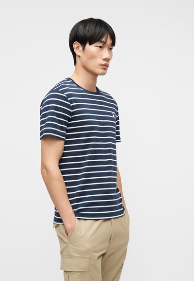 Νέος άνδρας που φοράει μπλε-σκούρο και λευκό ριγέ μπλουζάκι και μπεζ παντελόνι τύπου cargo με τα χέρια στις τσέπες, στέκεται μπροστά σε απλό φόντο.