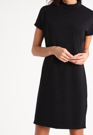 Žena nosící černé šaty s krátkým rukávem sahající po kolena a vysokým výstřihem, stojící před čistě bílým pozadím.