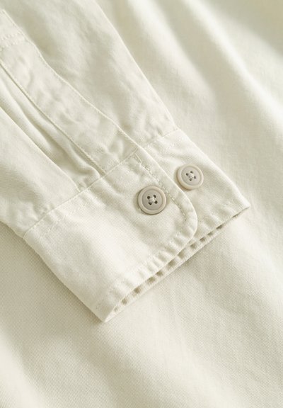 Gros plan sur un poignet de chemise beige avec deux petits boutons beige et des coutures détaillées sur du tissu en coton.
